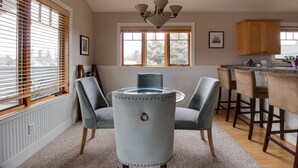 Un coin repas comprenant une table ronde en verre, quatre chaises grises à revêtement, un lustre, des meubles en bois et une fenêtre avec des stores.