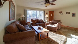 A living room with a brown leather sofa set, a wooden coffee table, a ceiling fan, and a large window providing natural light. - Cozy Tollgate Cabin Near Trails + Pool (Sisters)