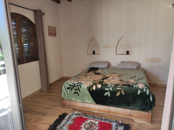 A bedroom with a wooden bed, floral bedspread, two pillows, a small mirror, and a window with curtains.