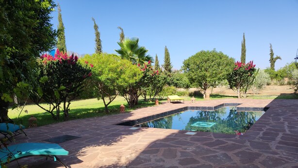 A stone-paved outdoor area with a swimming pool, surrounded by lush greenery and trees. - Maison Avec Piscine Privée, Jardin Arboré, Parking Clos à 2 kms D'amizmiz (Amizmiz)