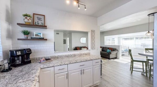 A modern kitchen with granite countertops, white cabinetry, and a coffee maker. There is a dining area with a table and chairs, and a living room with a sofa and a large window.