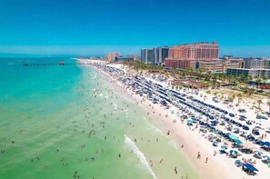 Ein Strand mit vielen Sonnenschirmen und Personen, einer Pier, die sich ins Wasser erstreckt, und mehreren Hotels im Hintergrund.