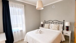 A neatly arranged bedroom with a bed, bedside tables, lamps, and a window with curtains. - Villa Ancor (Playa Blanca)