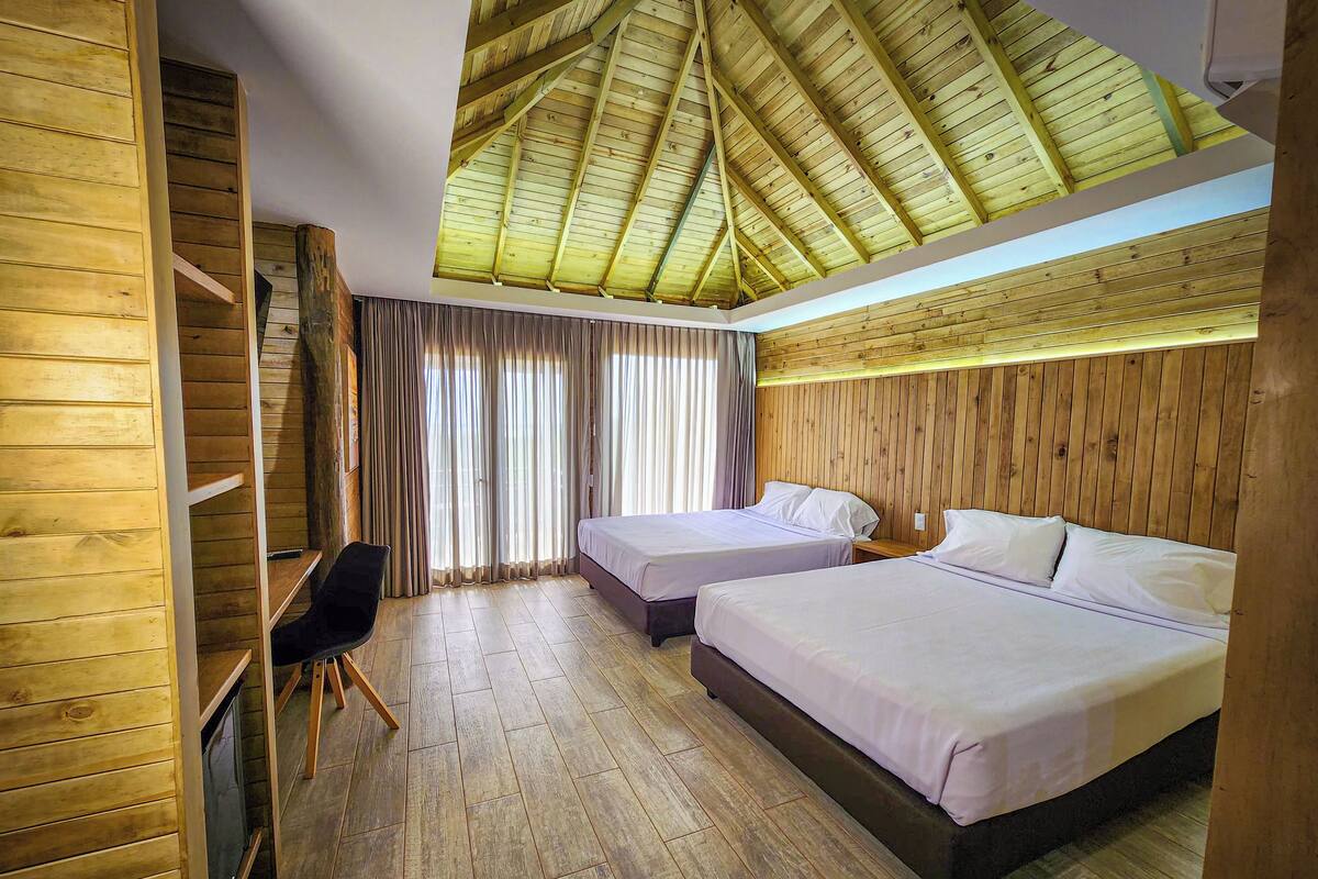 A room with two beds, wooden walls, a ceiling with wooden beams, and a desk with a chair.