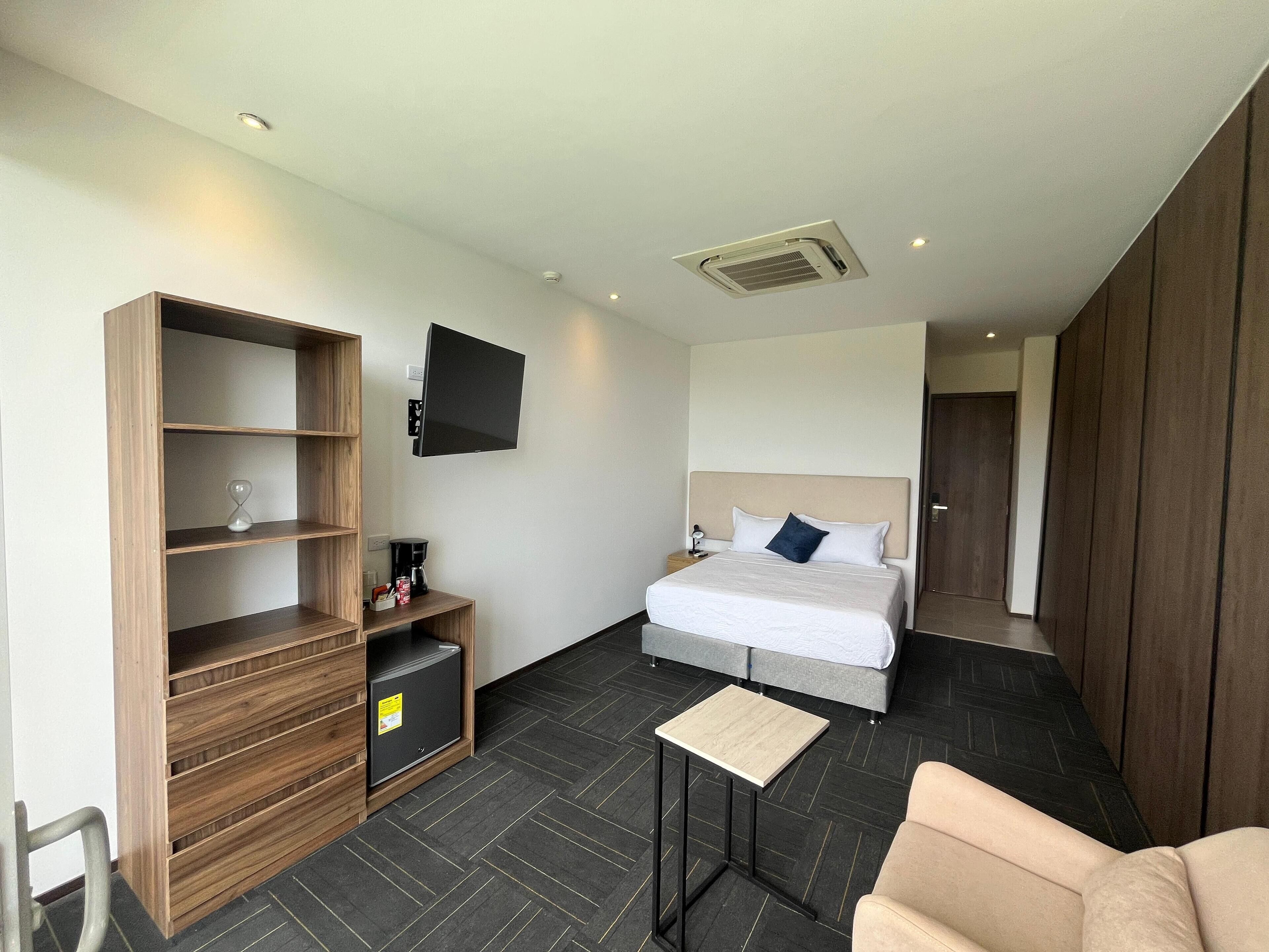 A modern hotel room with a bed, a TV mounted on the wall, a wooden shelf unit, a small table, and a chair.