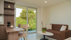 A modern living room with a brown sofa, a round coffee table, and a wooden dining table with chairs. There is a large sliding glass door leading to a balcony with a view of greenery.