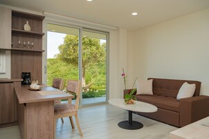 Una sala de estar moderna con un sofá marrón, una mesa de centro redonda y una mesa de comedor de madera con sillas. Hay un amplio ventanal corredera que da a un balcón con vistas a zonas verdes.