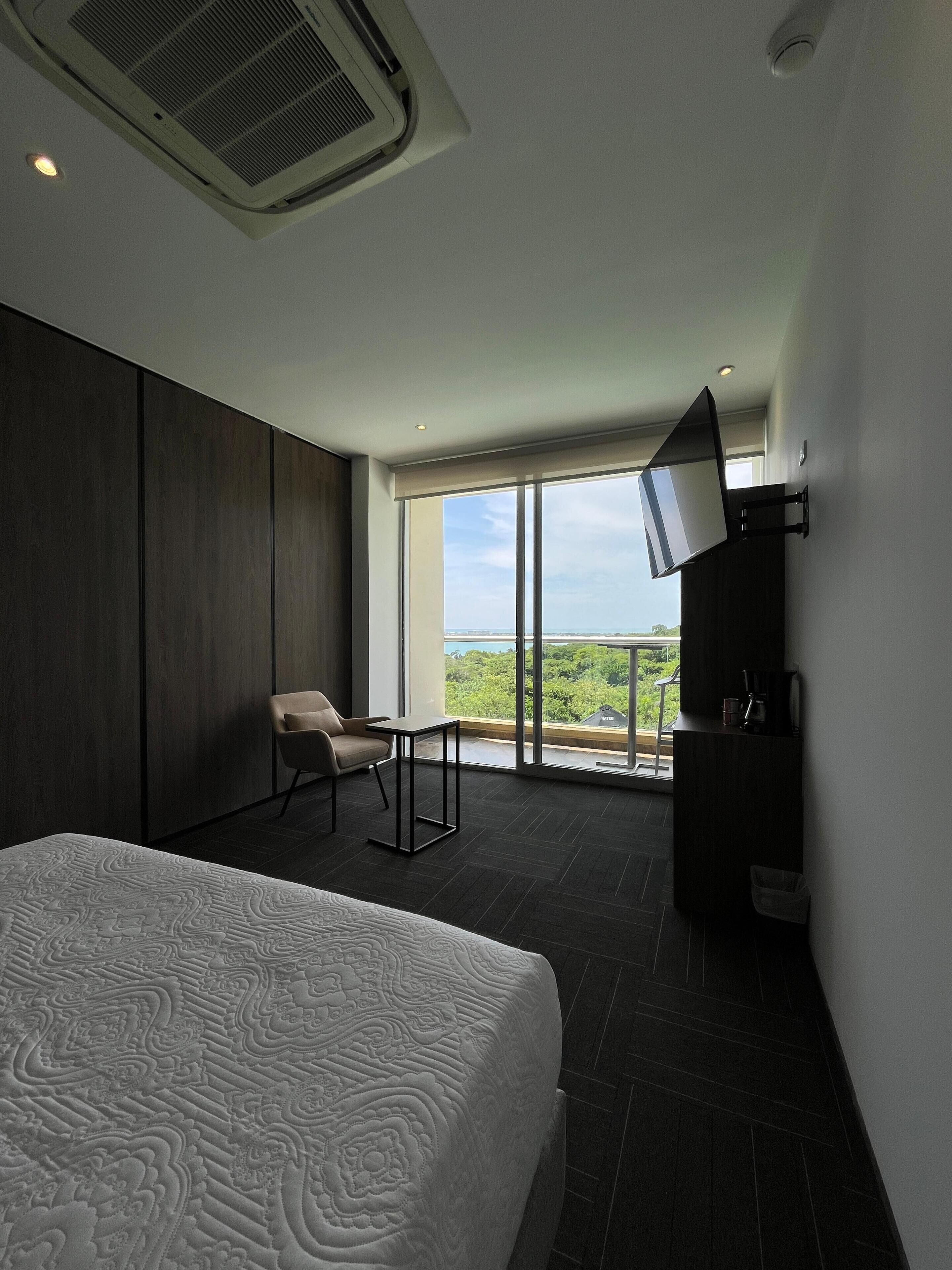 A modern hotel room with a large bed, a desk, a chair, a TV mounted on the wall, and a balcony with a view.