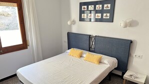 Un lit bien fait, avec deux oreillers, une petite table de chevet sur laquelle repose un magazine, et un cadre photo fixé au mur contenant plusieurs images.