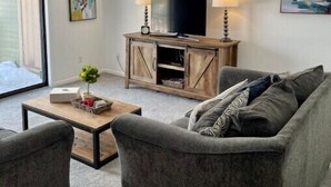 Ein Wohnzimmer mit grauem Sofa, einem Holz-TV-Schrank und einem Couchtisch. An den Wänden befinden sich zwei Wandleuchten und abstrakte Kunstwerke.