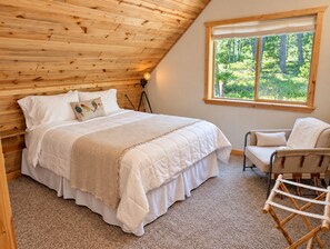 Un dormitorio con techo de madera, una cama grande con ropa de cama blanca, un ventanal con vistas a la vegetación y una silla con cojín blanco.