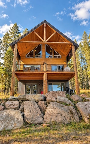 A two-story log cabin with a balcony, surrounded by large rocks and trees.