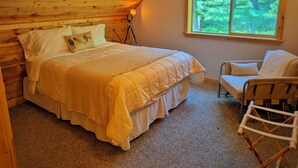 Quarto com teto de madeira, cama com colcha, cadeira e janela com vista para árvores.