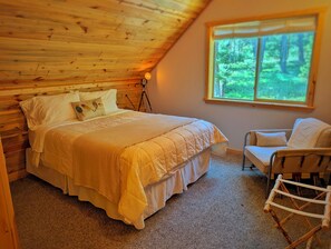 Una camera da letto con soffitto in legno, un letto con copriletto, una sedia e una finestra vista alberi.