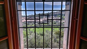Quarto com uma janela grande com grades de metal, piso de madeira e vista para uma paisagem verde com edifícios ao longe.