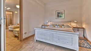 Quarto com cama branca, mesas de cabeceira, um criado-mudo, um espelho e uma porta para outro cômodo.