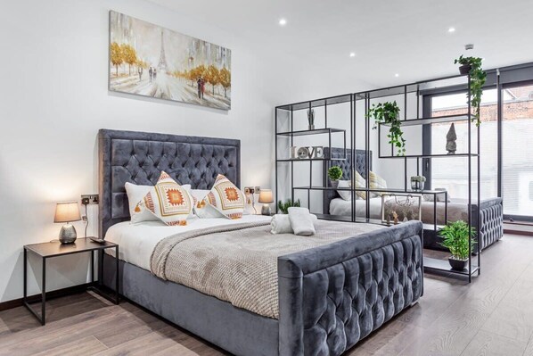 A modern bedroom with a large bed, a bedside table, a nightstand, a decorative shelf with plants, and a painting on the wall. - Luxury Sleeps 4 Studio Flat (Greater London)