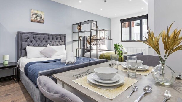 A modern bedroom with a tufted headboard bed, a dining area with a glass table set for two, and a large window providing natural light. - Serviced Studio Flat near Camden Euston Station (Greater London)