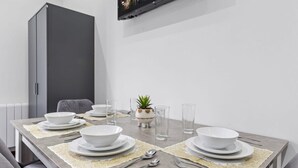 A modern dining room with a grey table set for four, including bowls, plates, cutlery, and glasses. A grey cabinet and a small potted plant are visible in the background.