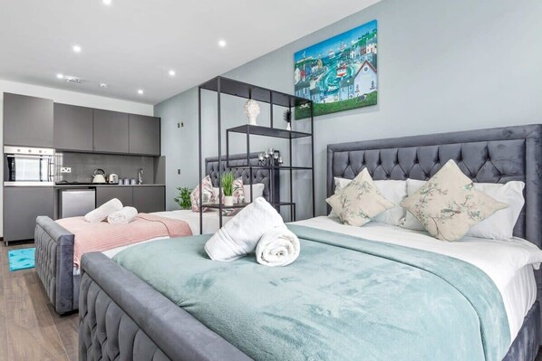 A modern hotel room with two beds, a kitchenette, and a cityscape painting on the wall.