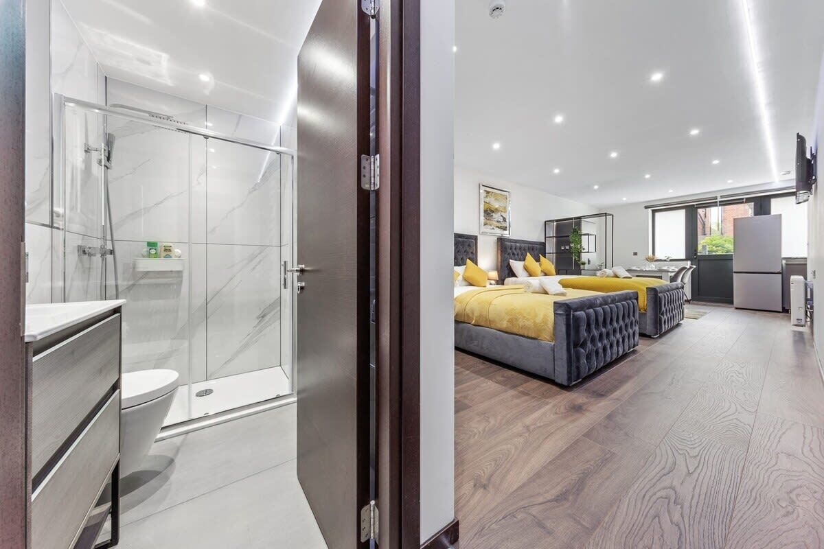 A modern hotel room with a glass shower enclosure, a bed with yellow and gray bedding, and a large window offering a view of the outdoors.