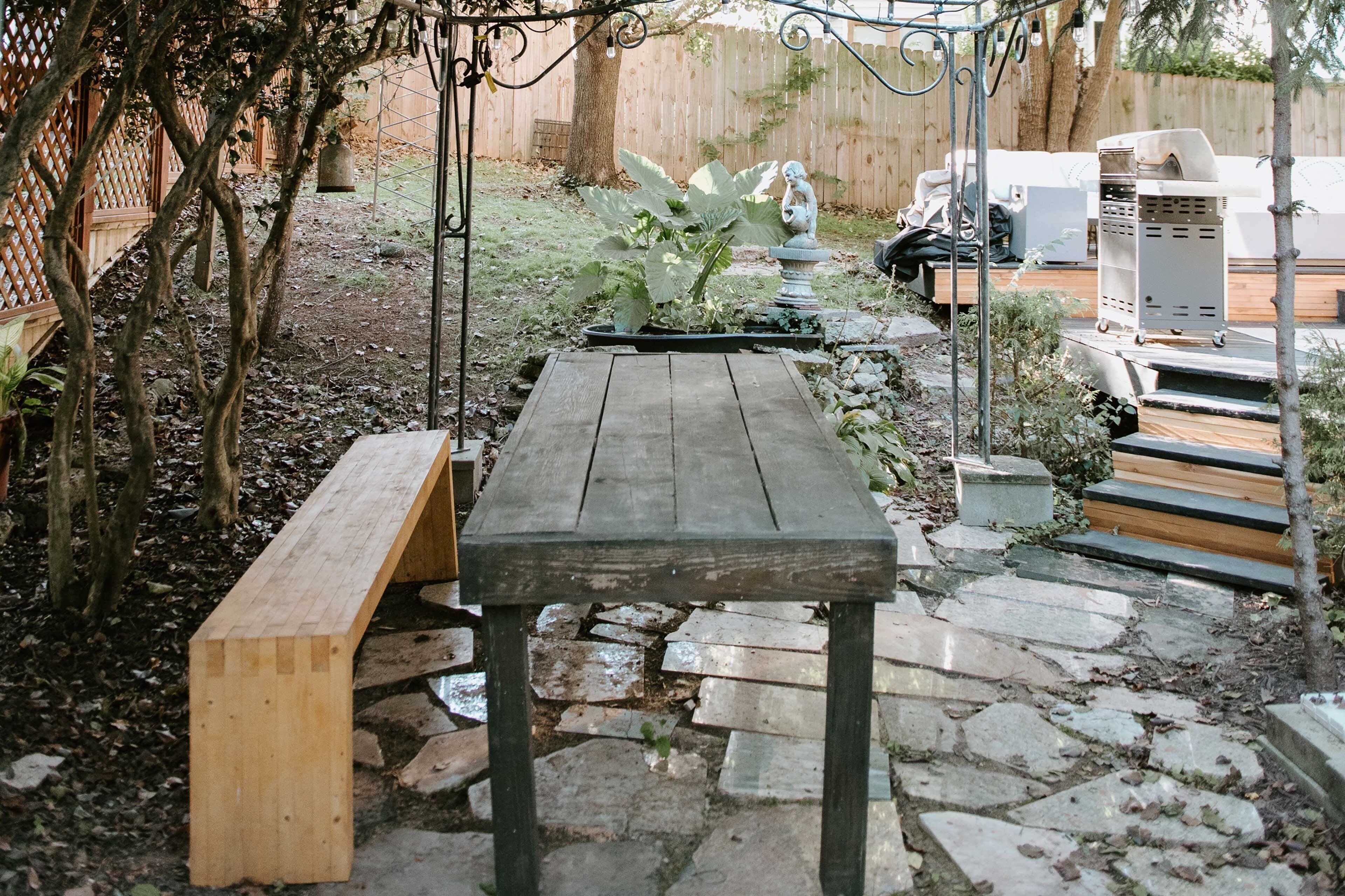 Un banc et une table en bois se trouvent dans un espace jardin, avec des allées pavées de pierres, le tout entouré de plantes et d’une clôture en bois.