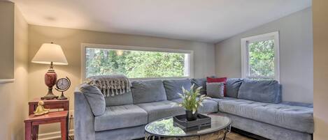 A living room with a grey sectional sofa, a glass coffee table, a lamp, and a small table with a clock and a decorative item.