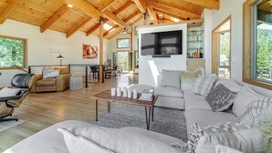 Un vaste espace de vie doté d’un haut plafond en bois, d’un grand téléviseur à écran plat fixé au mur et d’un coin salon confortable comprenant un canapé et un fauteuil.