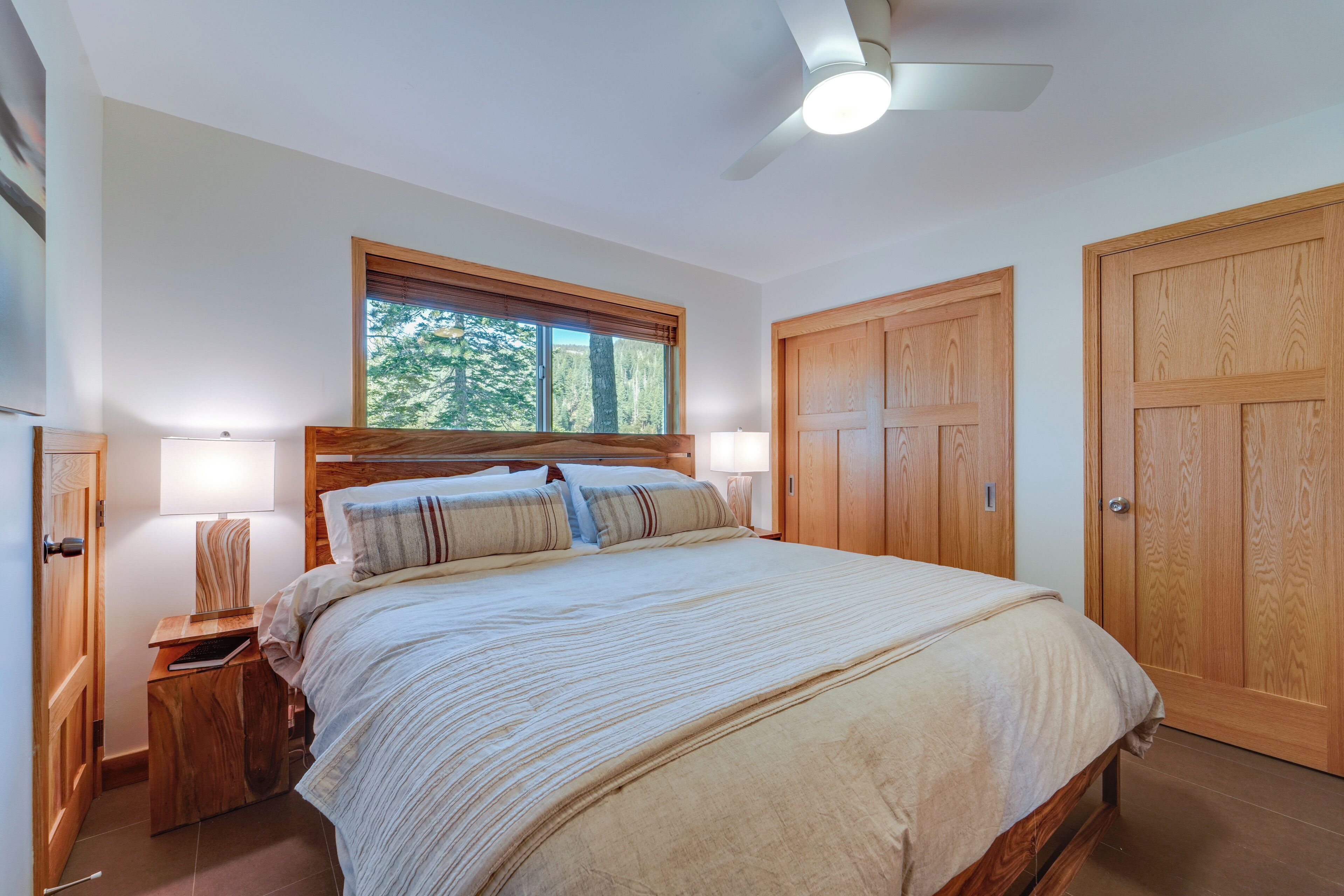 Un dormitorio con una cama grande, cabecera de madera, dos lámparas de noche, una mesita de noche, una ventana con persianas y un ventilador de techo.