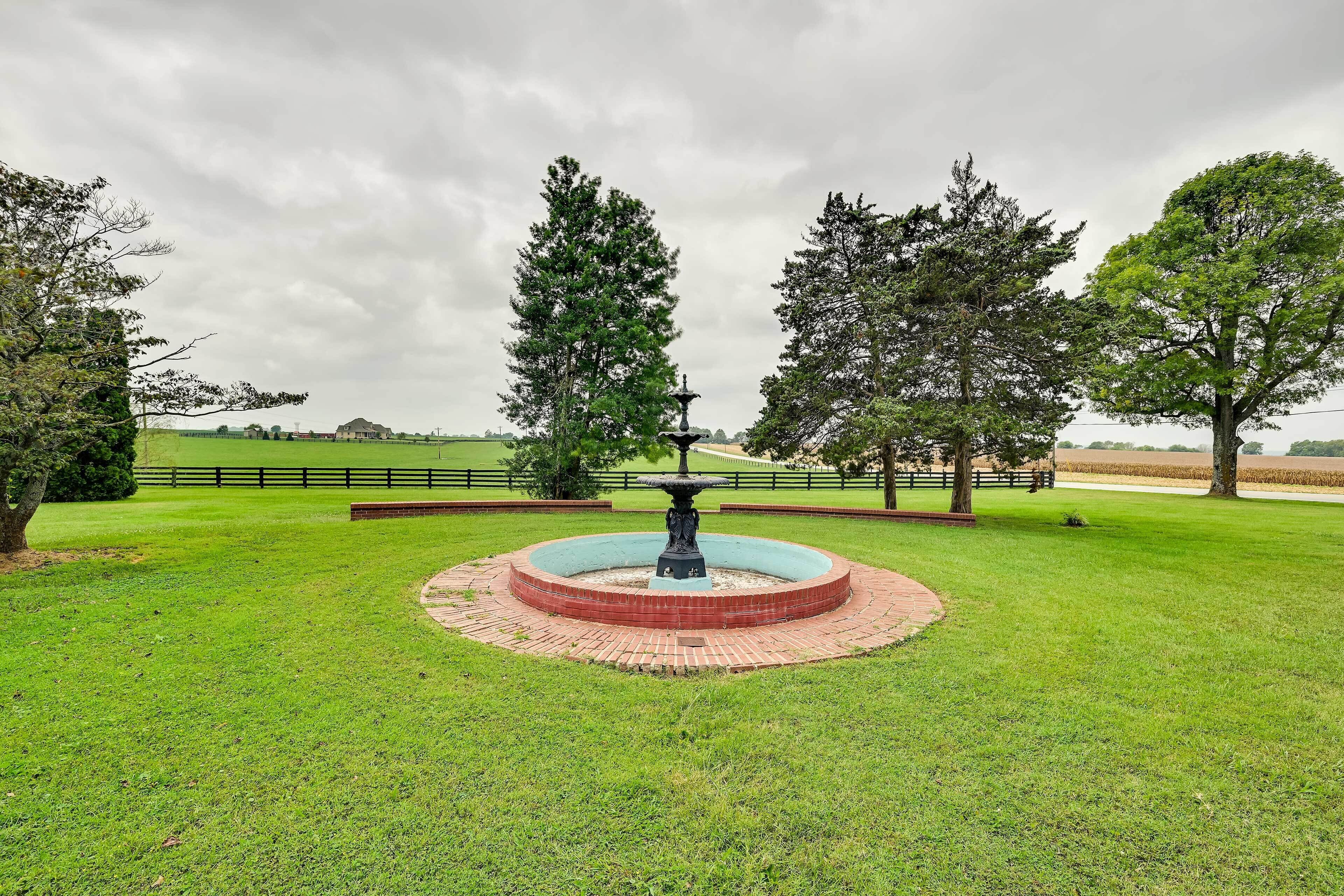Una fuente de ladrillo circular con una estatua en el centro, rodeada de un césped cuidado y altos árboles.