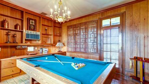 A pool table with a wooden backdrop, a chandelier, and a window with blinds. - Jennys Southland Homestead, LLC (Oakland)