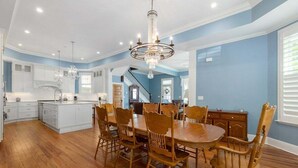 Un vaste espace dédié à la salle à manger, avec une grande table et des chaises en bois, un lustre et une cuisine en arrière-plan.