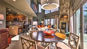 A spacious living area with a round wooden table, comfortable seating, a stone fireplace, and large windows offering a view of the outdoors. - Luxe Rumbling Bald Home w/ Deck & Mountain View (Lake Lure)