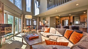 A spacious living area with a large sofa, a coffee table, and a television. There is a dining area with chairs and a table, and a kitchen with wooden cabinets and a countertop. - Luxe Rumbling Bald Home w/ Deck & Mountain View (Lake Lure)