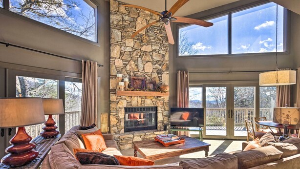 A spacious living room with a stone fireplace, large windows, and a ceiling fan. - Luxe Rumbling Bald Home w/ Deck & Mountain View (Lake Lure)