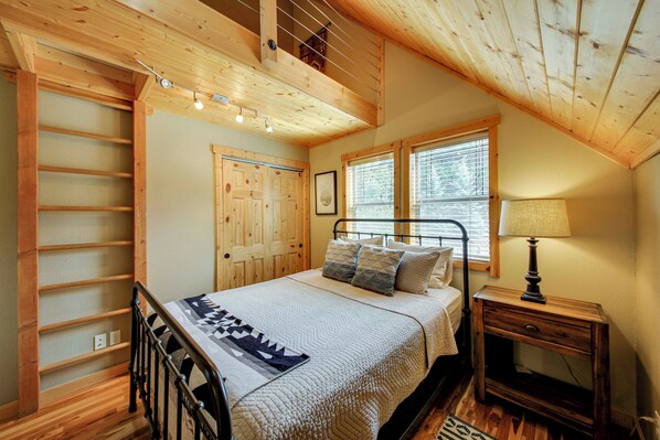 Une chambre à coucher avec un plafond en bois, un lit recouvert d’une courtepointe, une table de chevet et une fenêtre avec des stores.
