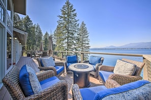 A deck with wicker furniture and a round table, overlooking a lake with trees and mountains in the background. - Lake Pend Oreille Waterfront With Amazing Views (Sagle)