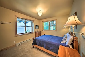 A bedroom with a large window, a bed with a blue quilt, a wooden nightstand, and a wall-mounted lamp. - Lake Pend Oreille Waterfront With Amazing Views (Sagle)