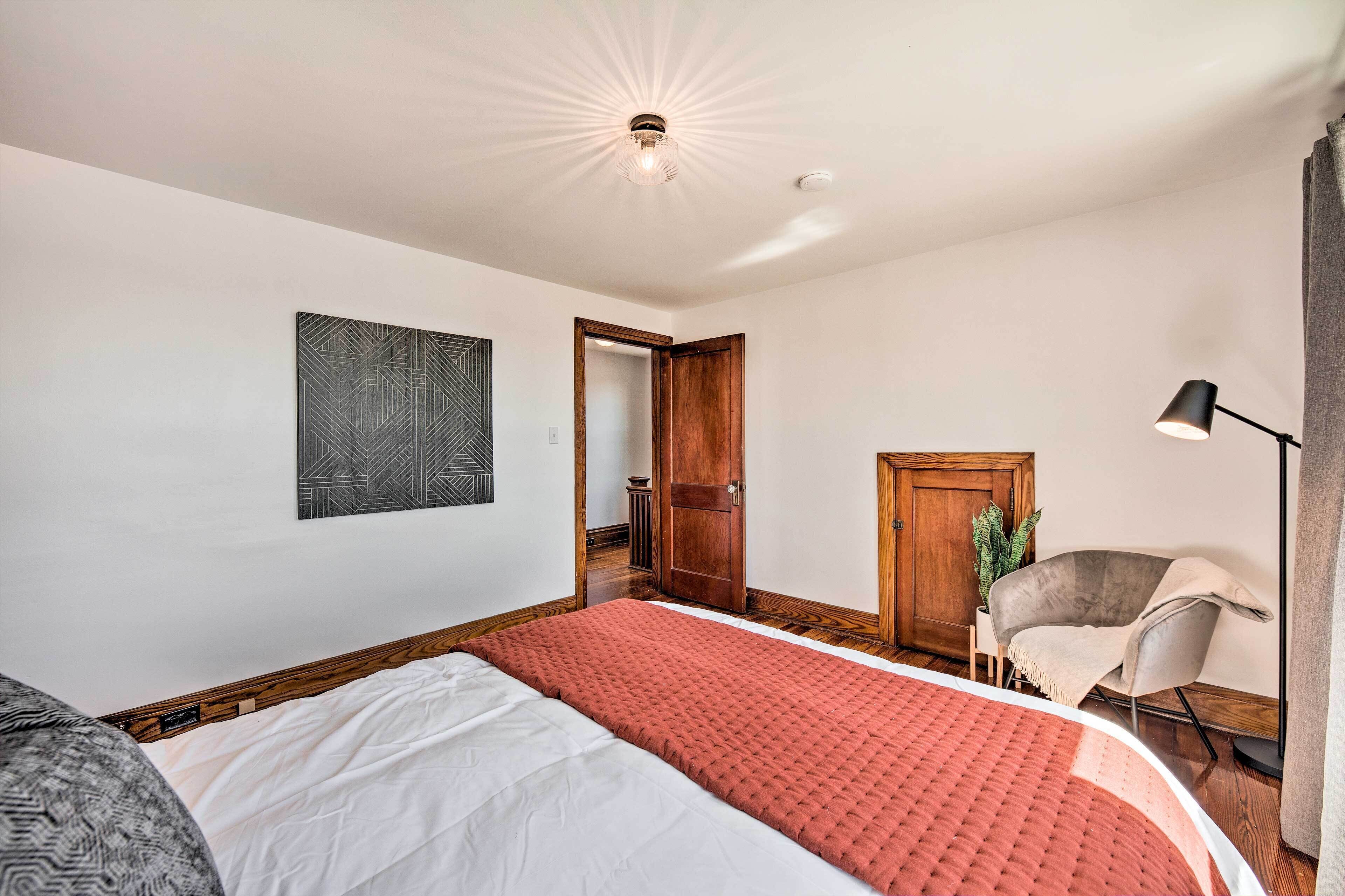 Une chambre à coucher avec un grand lit, une chaise, un bureau, une plante et une porte donnant sur une autre pièce.