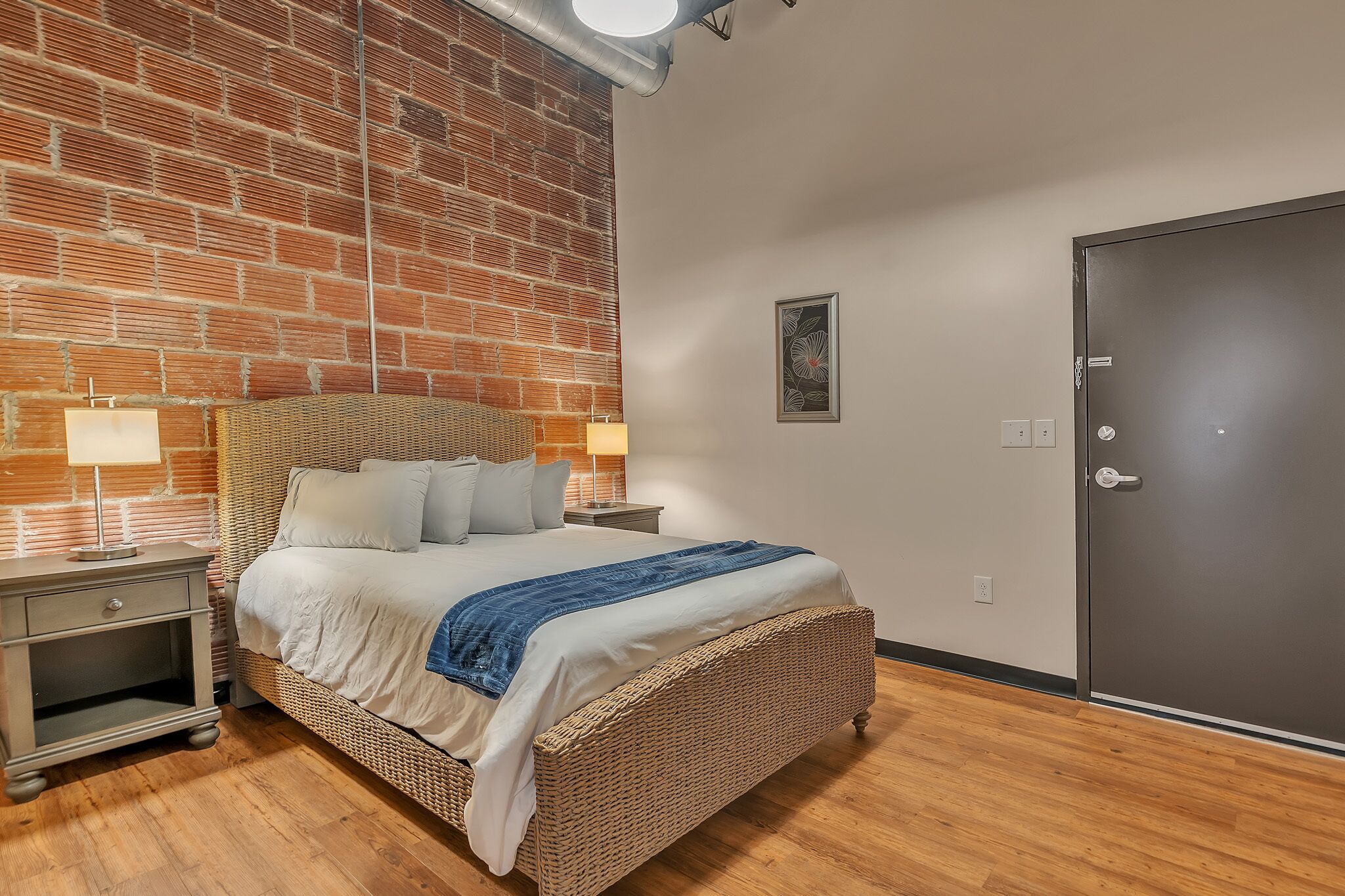 Une chambre avec un mur en briques, un lit avec une literie blanche et un plaid bleu, une table de chevet avec une lampe, et une porte.