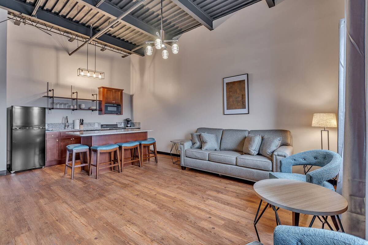 A modern kitchen with a central island, bar stools, a grey sofa, and a round table.