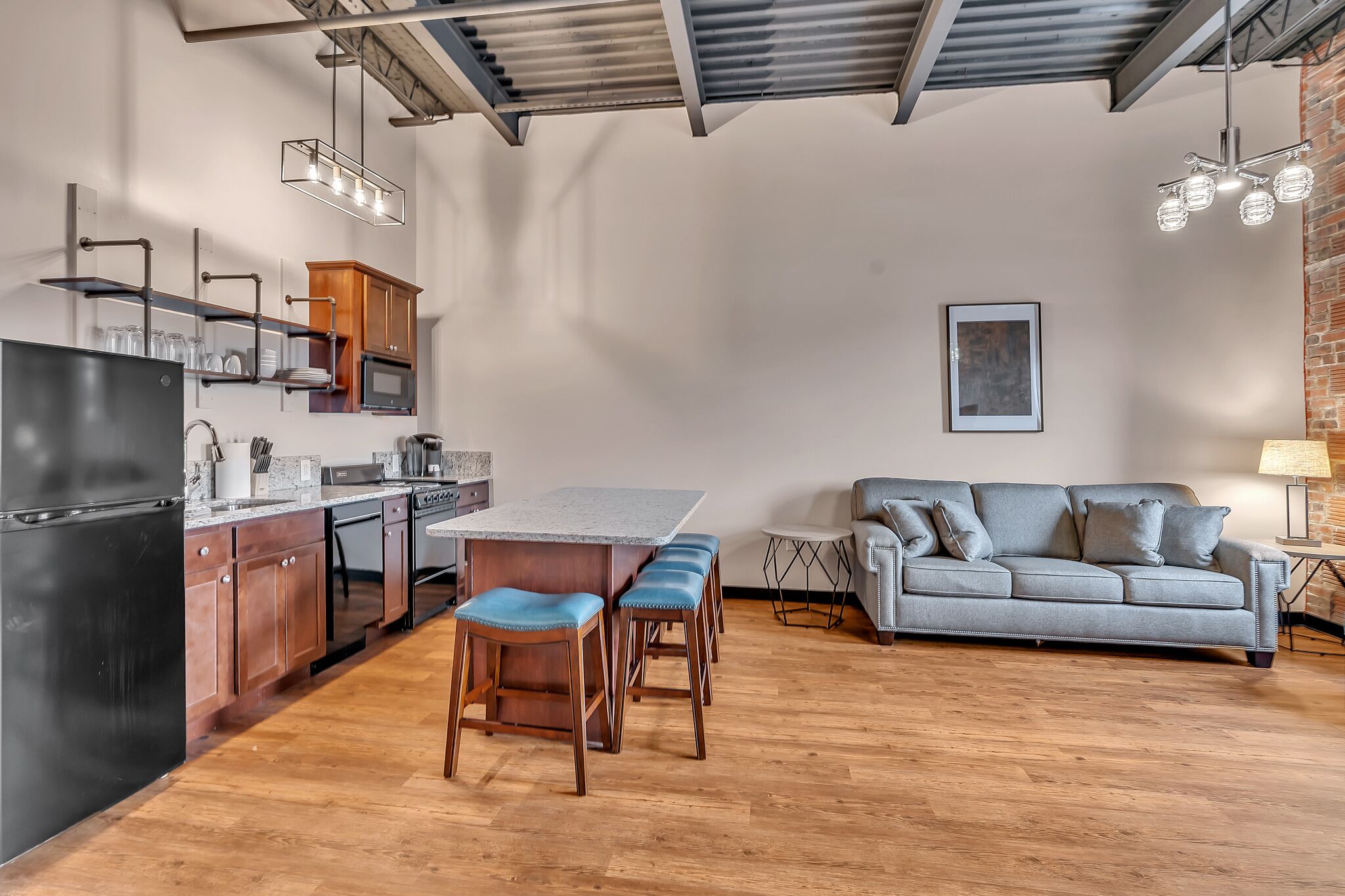 A modern kitchen with a central island, wooden bar stools, and a grey sofa in a living area.