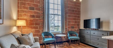 A living room with a brick wall, a large window, a sofa, two chairs, a TV, and a cabinet.