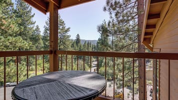 Una terraza de madera con una mesa redonda, rodeada de barandas metálicas, con vistas a una zona boscosa de pinos y un cielo despejado.