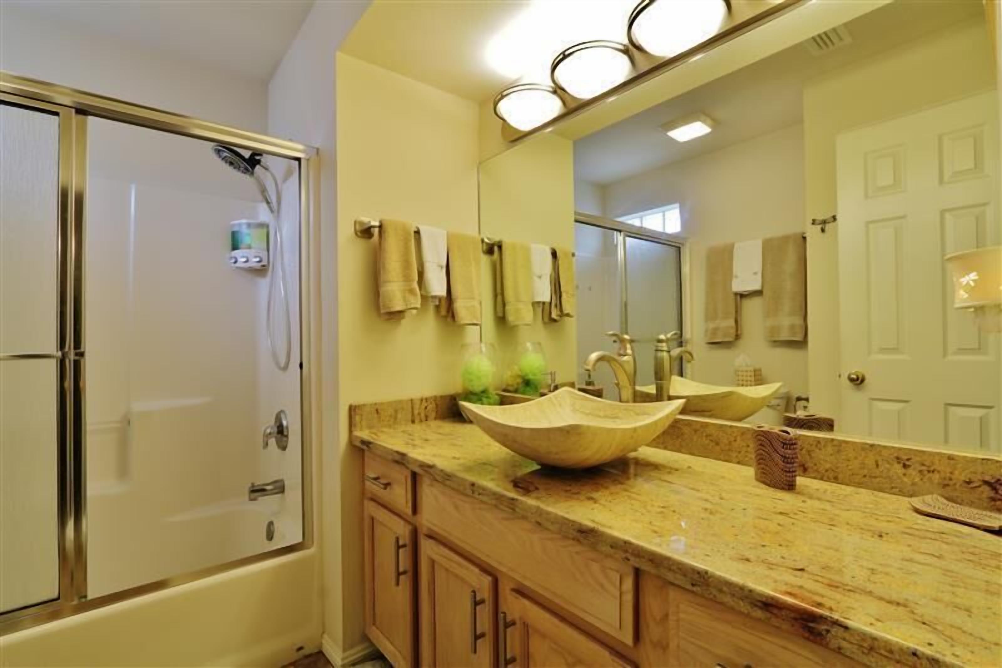 A bathroom with a large mirror, a wooden vanity, a beige sink, and a shower.
