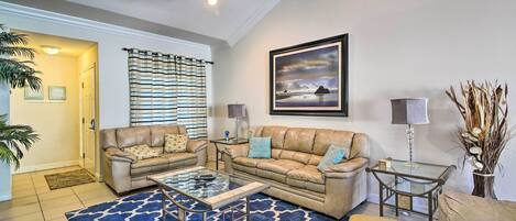A living room with a beige sofa set, a glass coffee table, a striped curtain, a framed landscape painting, and a vase with dried plants.