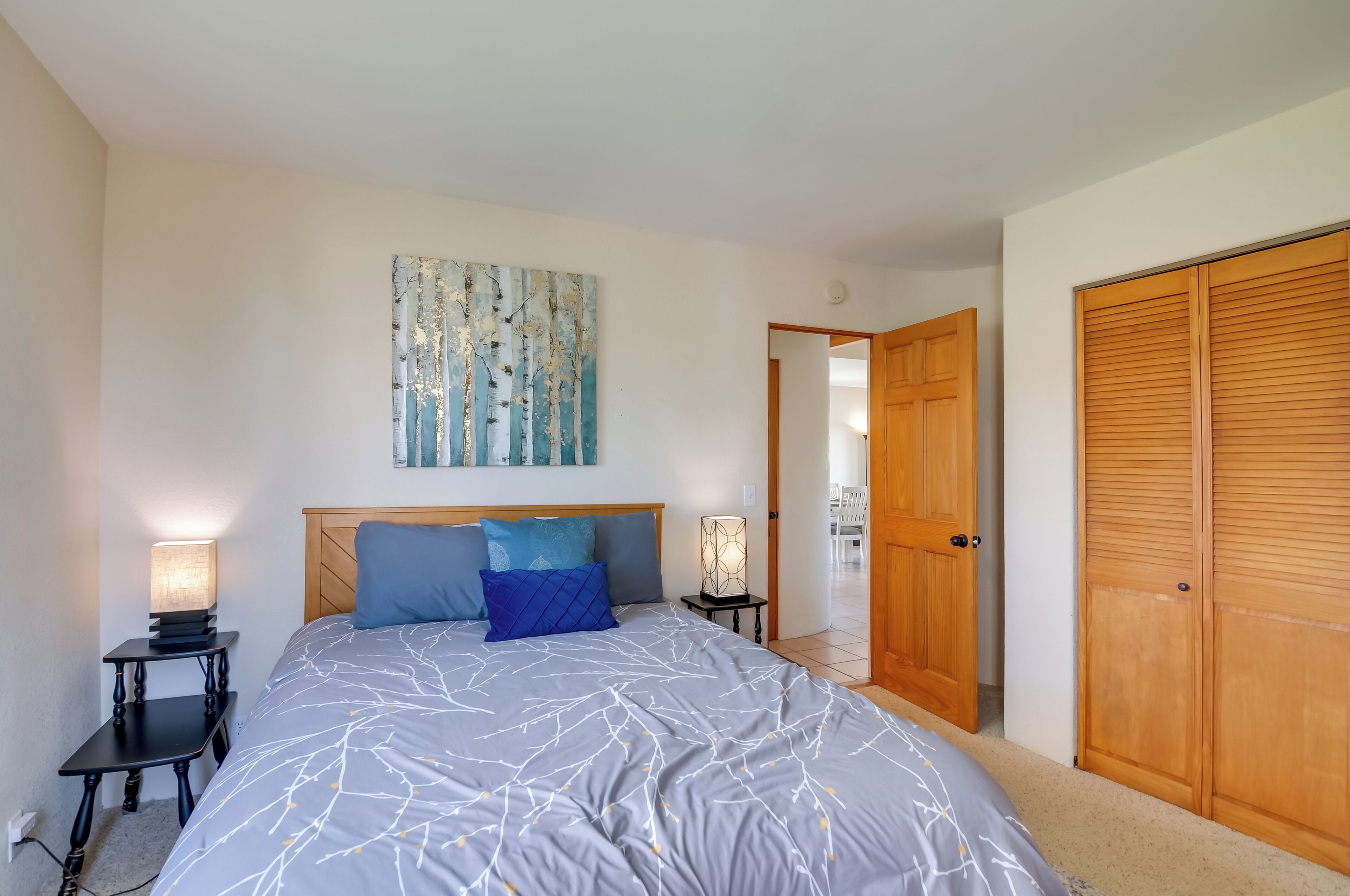 Un dormitorio con una cama grande, dos mesitas de noche con lámparas, un armario de madera y una puerta que da a otra habitación.