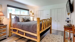 Quarto com cama de madeira, mesa de cabeceira, luminária, televisão fixada na parede e janela com persianas.