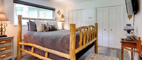 A bedroom with a wooden bed, a bedside table, a lamp, a TV mounted on the wall, and a window with blinds.