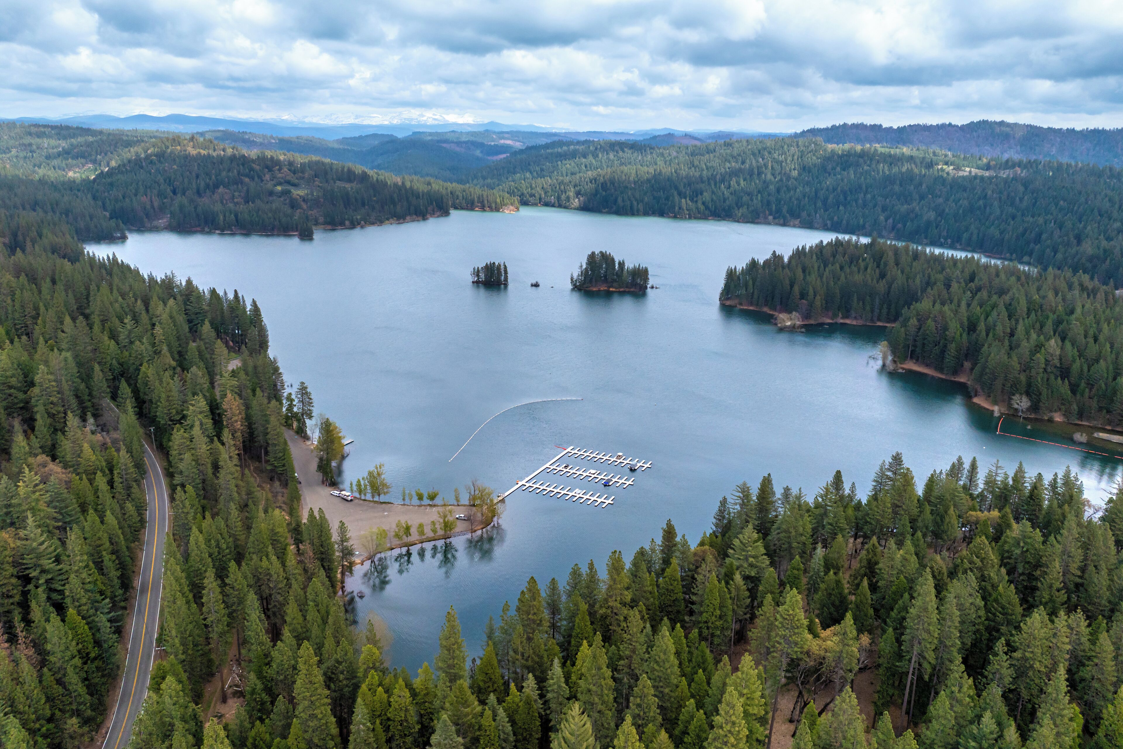 Ein See, von dichtem Wald umgeben, eine durch die Bäume führende Straße und ein Steg, der ins Wasser ragt.